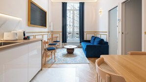 A modern hotel room with a kitchen area, a dining table, a blue sofa, and a view of the city through large windows. - Parisian Charm by the Louvres (Paris)