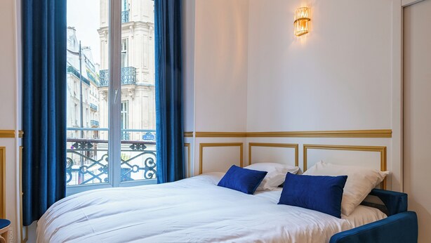 A hotel room with a large bed, a view of the city through a balcony, and a wall-mounted light fixture. - Parisian Charm by the Louvres (Paris)