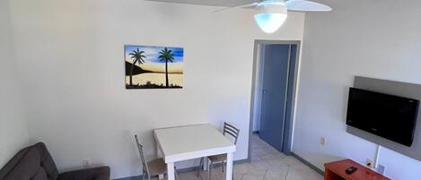 A living room with a sofa, a dining table with chairs, a ceiling fan, a painting, a TV, and a small wooden table.
