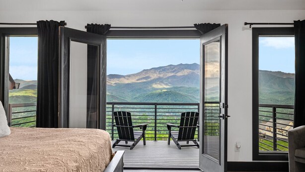 A bedroom with a large bed, a balcony with two chairs, and a view of mountains. - Swim Spa/VIEWS/Hot Tub/Game Room - Viewfinder (Gatlinburg)