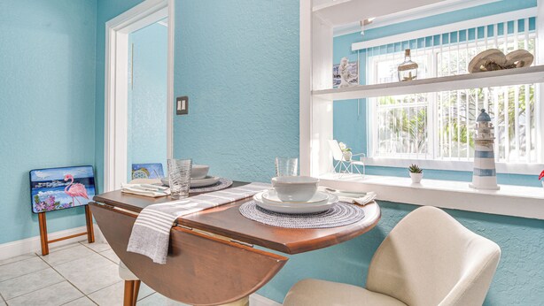 A dining area with a table set for two, a chair, a window with decorative items, and a painting on the wall. - Walk to Dining & Sand! St Pete Beach Apartment (St Pete Beach)