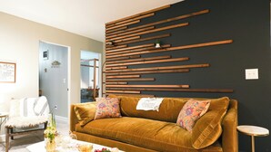Un salon avec un canapé de couleur moutarde, une table basse blanche sur laquelle sont disposés des en-cas et un panneau en bois fixé au mur.