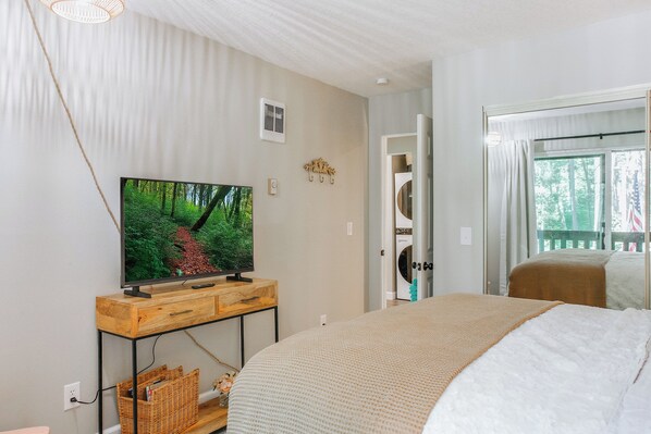 A bedroom with a large bed, a television on a wooden stand, a wicker basket, and a view of a balcony with curtains.