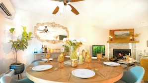 Une salle à manger avec une table ronde dressée pour quatre personnes, entourée de chaises bleues. On aperçoit une cheminée au fond de la pièce, et un grand miroir est accroché au-dessus de la table.