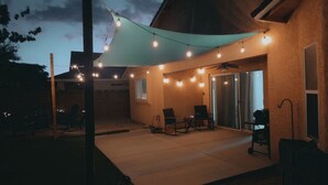 Un patio al aire libre con luces colgantes, una zona de estar cubierta y un ventanal corredero que da a un garaje techado.