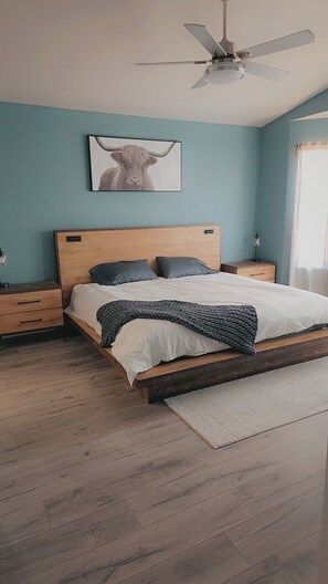 A bedroom with a wooden bed, a ceiling fan, a nightstand, a bedside table, and a wall-mounted picture of a bull. - Chateau Relaxo (Kingman)