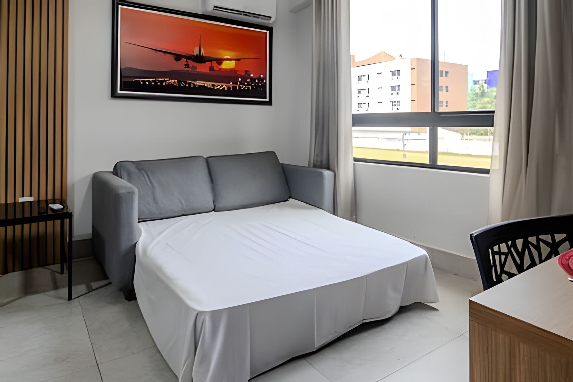 Ein Hotelzimmer mit Bett, Schreibtisch, Stuhl, Fenster mit Blick auf Gebäude und einem am Wand hängenden Bild von einem Flugzeug.