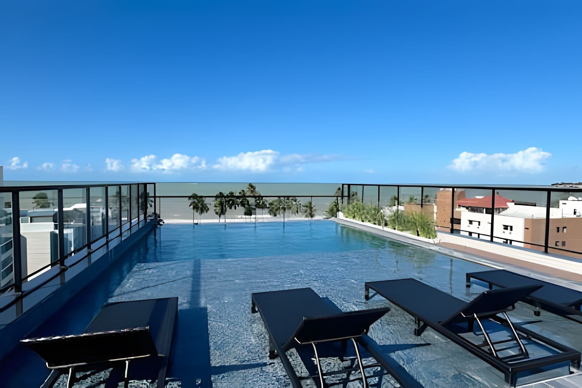A rooftop infinity pool with lounge chairs, clear skies, and a view of the beach.