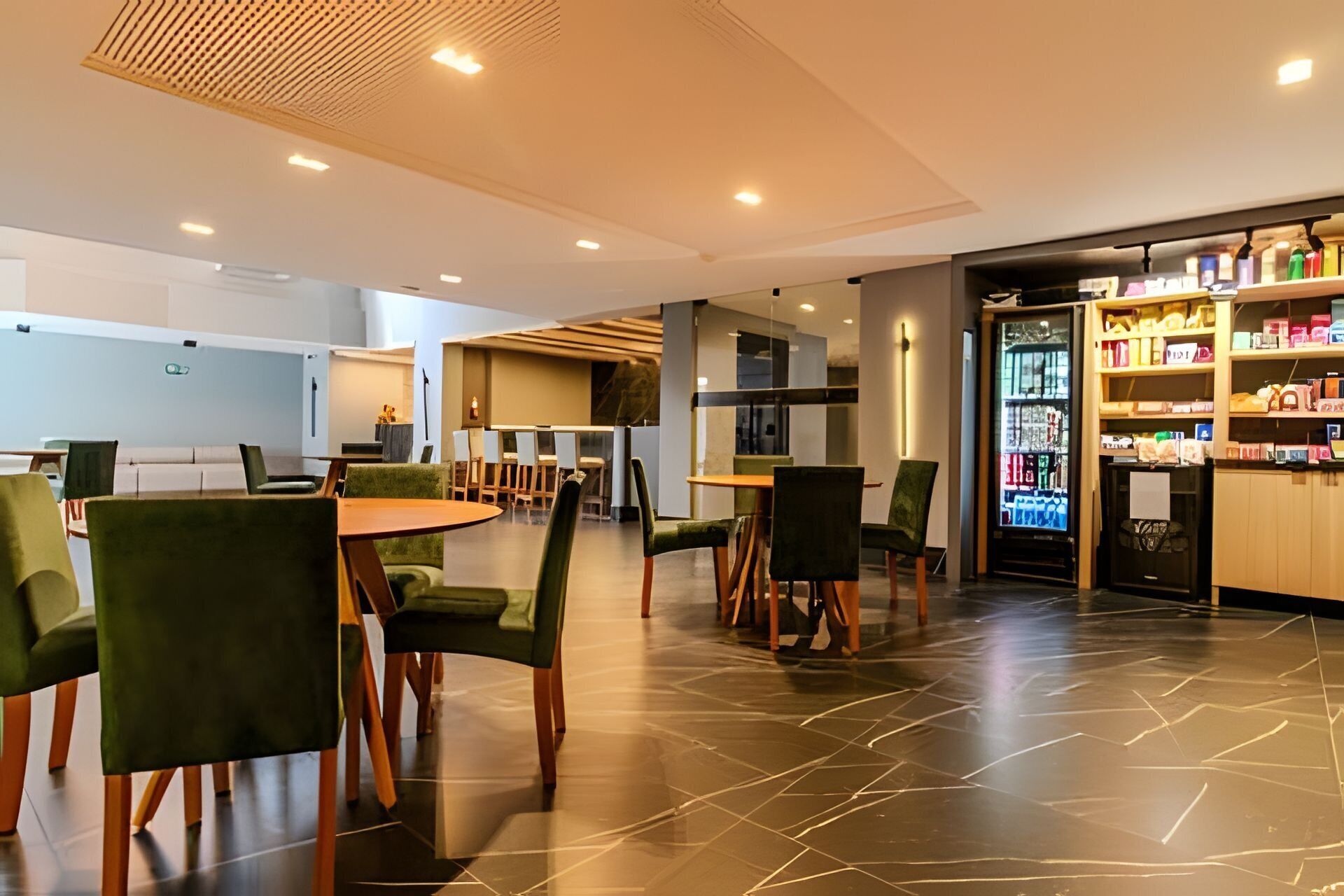 A modern dining area with green upholstered chairs, a round table, and a vending machine stocked with beverages.