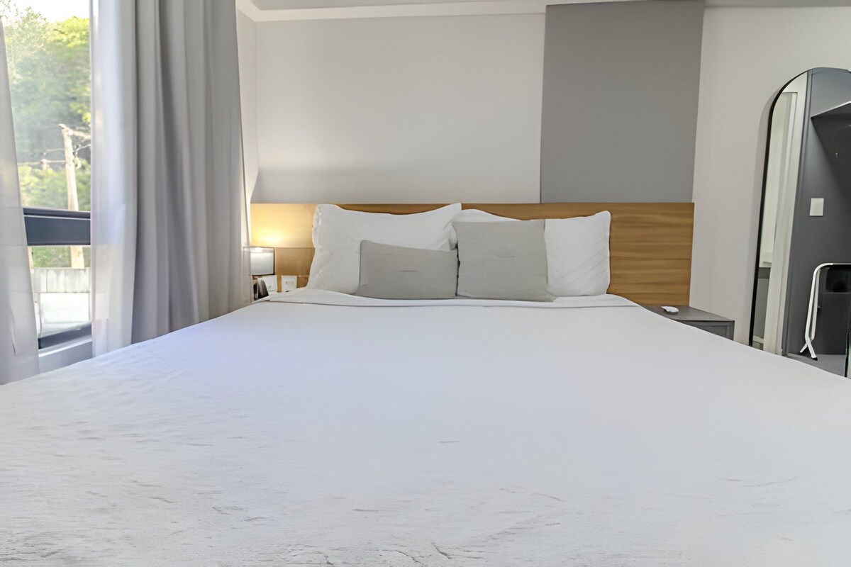A hotel room with a large bed, a wooden headboard, a nightstand, a lamp, and a view of the outdoors.