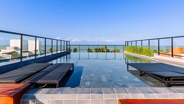 A rooftop infinity pool with clear views of the ocean and palm trees, surrounded by glass railings and sun loungers. - Legacy Suites (João Pessoa)