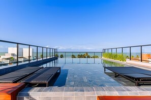 A rooftop infinity pool with clear views of the ocean and palm trees, surrounded by glass railings and sun loungers. - Legacy Suites (João Pessoa)