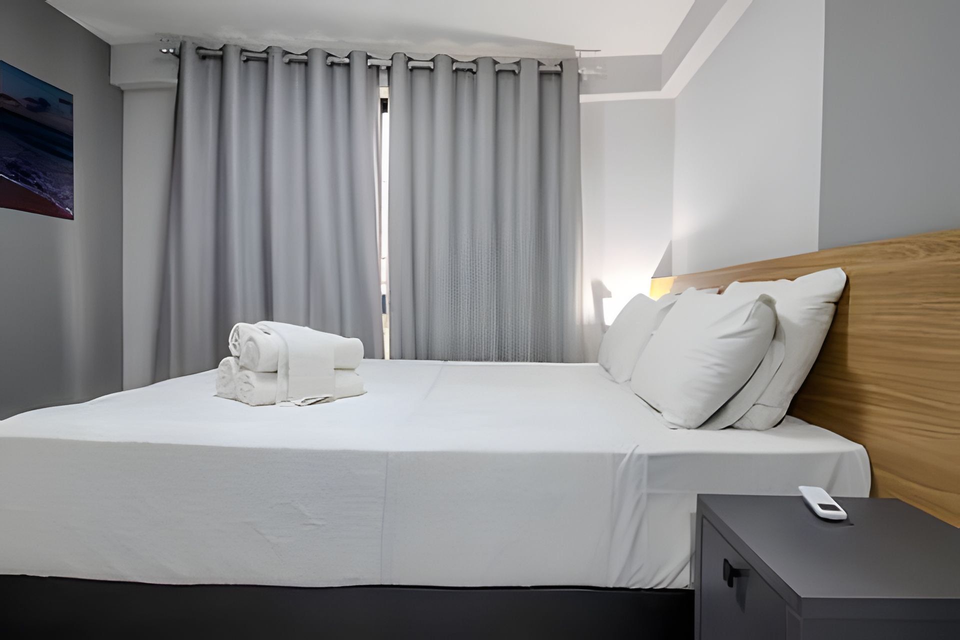 A hotel room with a bed, a nightstand, a remote control, and a window with curtains.