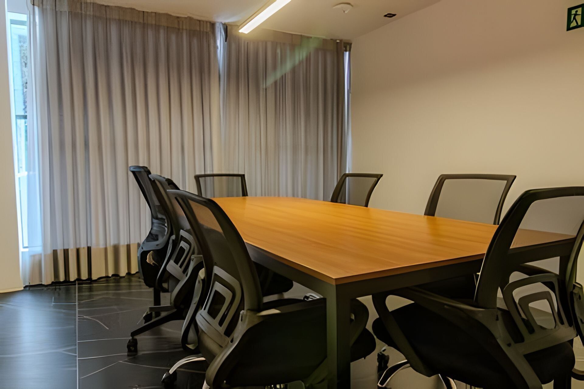 Ein Konferenzraum mit Holztisch und schwarzen Stühlen, großen Fenstern mit Vorhängen sowie einer schlichten Wand mit einem Brandschutztürschild.