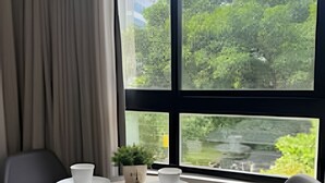 A room with a table, two chairs, a window with a view of greenery and a building, and a hat on one chair. - Legacy Suites (João Pessoa)
