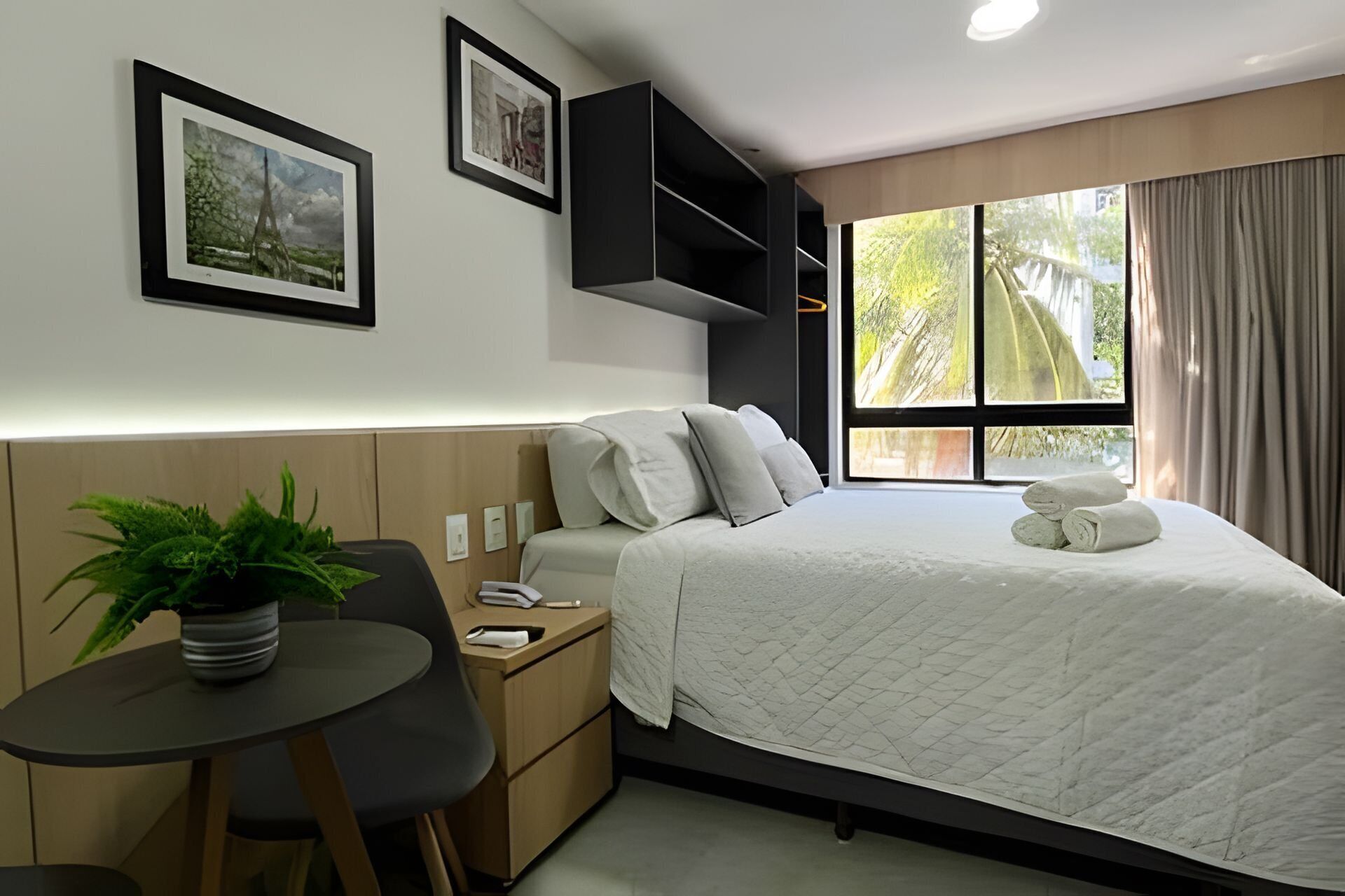 A hotel room with a bed, a desk, a chair, a potted plant, and a window with curtains.