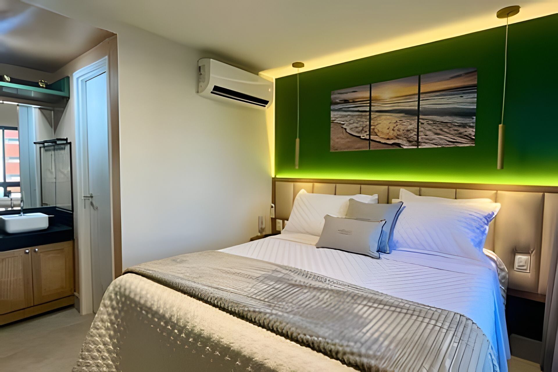 Habitación de hotel con cama, mesita de noche, aire acondicionado de pared, baño con lavamanos y pared verde con un cuadro.
