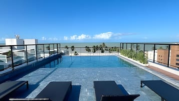 Una piscina en la azotea con agua azul cristalina, sillones de descanso y vistas al océano y edificios a lo lejos.