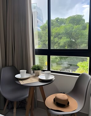 A room with a table, two chairs, a window with a view of greenery and a building, and a hat on one chair. - Legacy Suites (João Pessoa)