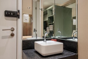 A bathroom with a white sink, two faucets, a mirror, and a door with a handle and lock. - Legacy Suites (João Pessoa)