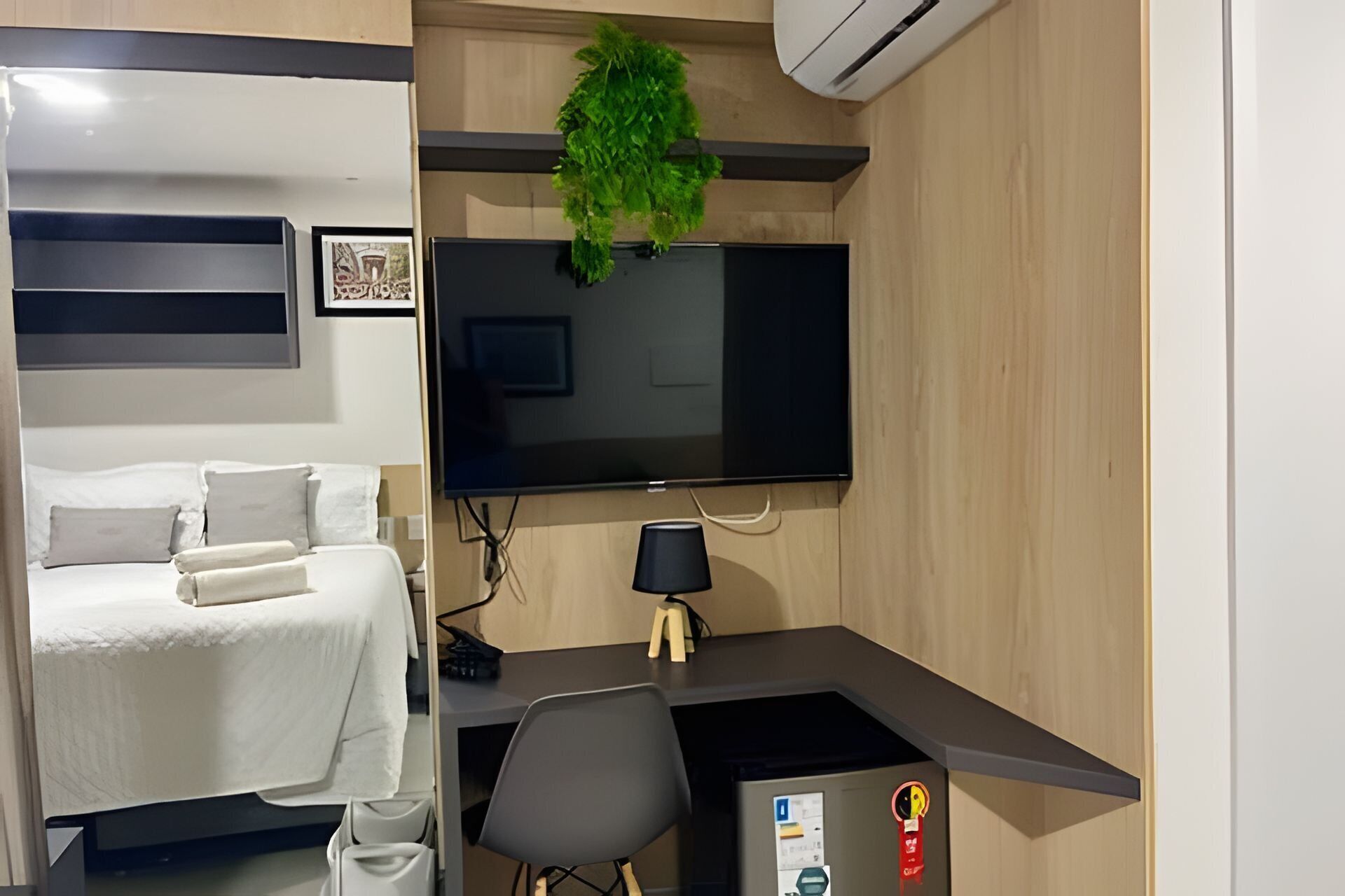 A compact hotel room with a bed, a desk with a chair, a flat-screen TV, a wall-mounted air conditioner, and a small plant.