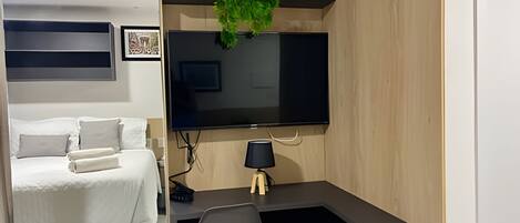 A compact hotel room with a bed, a desk with a chair, a flat-screen TV, a wall-mounted air conditioner, and a small plant.