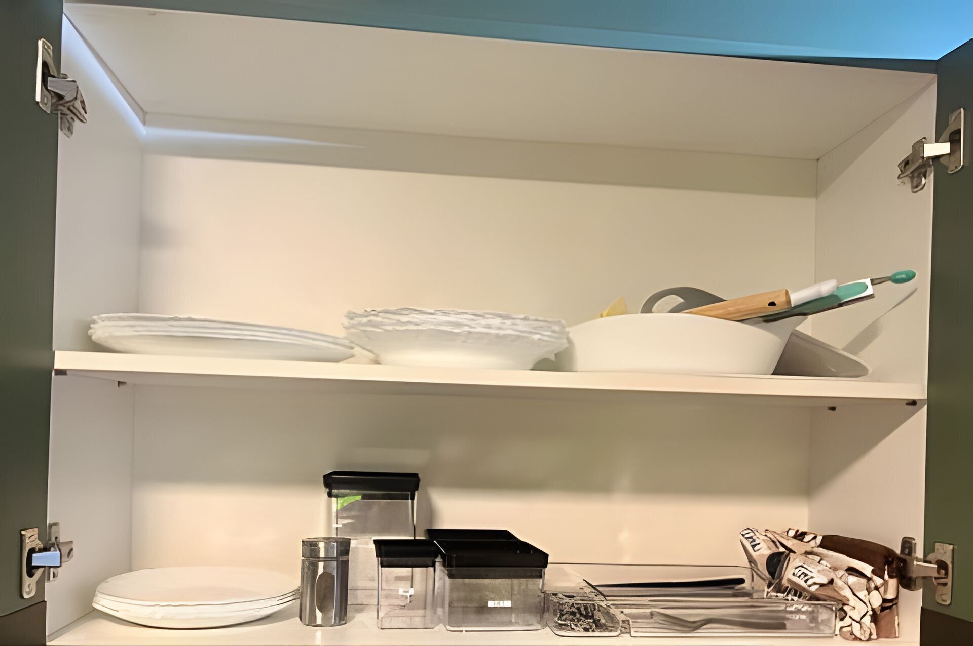 A kitchen cabinet with white shelves, containing stacked plates, bowls, a knife, and various containers.