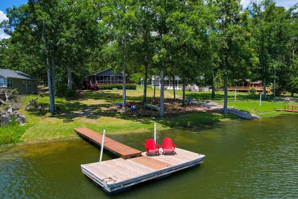 A dock with two red chairs, a grassy area, and a house in the background. - Easy Lake Living - Sleeps 8 (Prosperity)