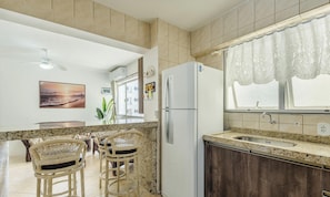 Een keuken met een witte koelkast, een granieten aanrechtblad en donkerhouten kasten. Er staan twee rieten barkrukken en een eettafel met stoelen.