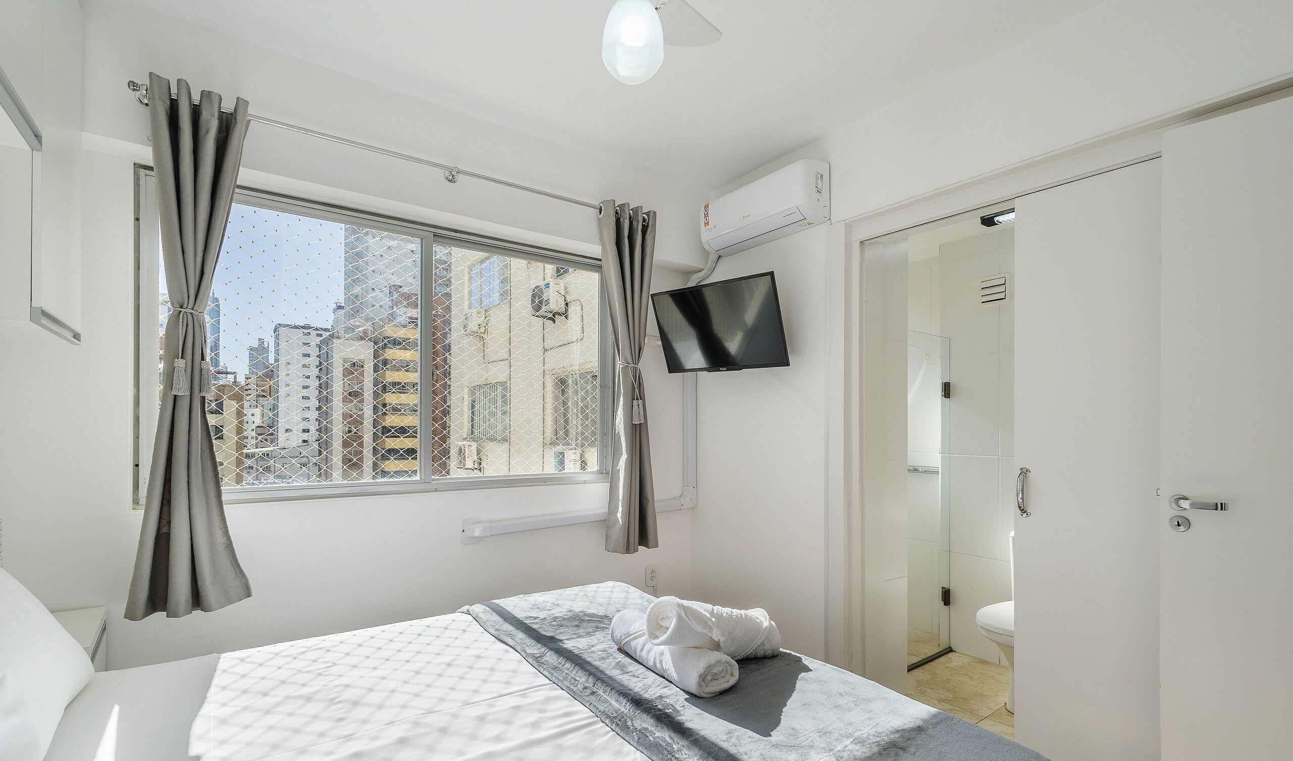 A hotel room with a bed, a TV mounted on the wall, a window with curtains, and a door leading to a bathroom.