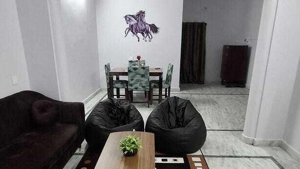 Una sala de estar con un sofá marrón oscuro, dos pufs negros, una mesa de centro de madera con una planta pequeña y una alfombra estampada.