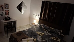Un dormitorio con cama, escritorio, silla, mesita de noche con lámpara y una obra de arte en la pared.