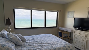 A bedroom with a bed, a TV, a desk, and a window with a view of the sea.