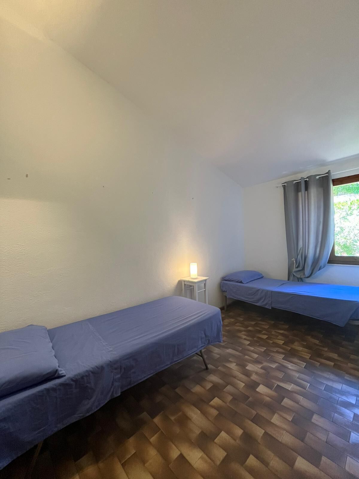 Eine kleine, einfache Unterkunft mit zwei Einzelbetten, einem Nachttisch mit Lampe und einem Vorhangfenster.