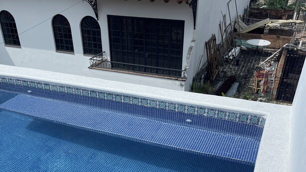 A rooftop swimming pool with blue mosaic tiles, a white wall, and a red-tiled roof.