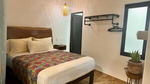 A bedroom with a bed, a bedside table, a hanging light fixture, a coat rack, a window, and a potted plant.