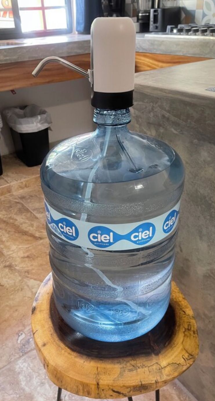 A large water dispenser with a white top and a blue label, placed on a wooden stool in a kitchen setting.