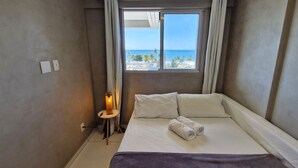 Une chambre d’hôtel avec un lit, une table de chevet, une lampe et une fenêtre donnant sur la plage et l’océan.