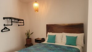 Ein ordentlich bezogenes Bett mit Holz-Kopfteil, einer hängenden Deckenleuchte, einem an der Wand befestigten Kleiderhaken und einer Blumentopf-Pflanze.
