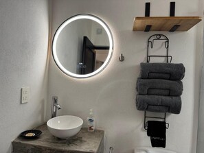 A bathroom with a round mirror, a white sink, a soap dispenser, and a towel rack with towels. - Spanish Moon PV - #14 Studio Queen (Puerto Vallarta)