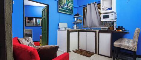 Un espacio habitable compacto con kitchenette, un sofá rojo, un sillón estampado y una pared azul.
