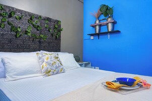 Een net opgemaakt bed met witte lakens en decoratieve kussens, een blauwe muur met een plank waarop decoratieve objecten staan, en een dienblad met een kom en servetje op het nachtkastje.