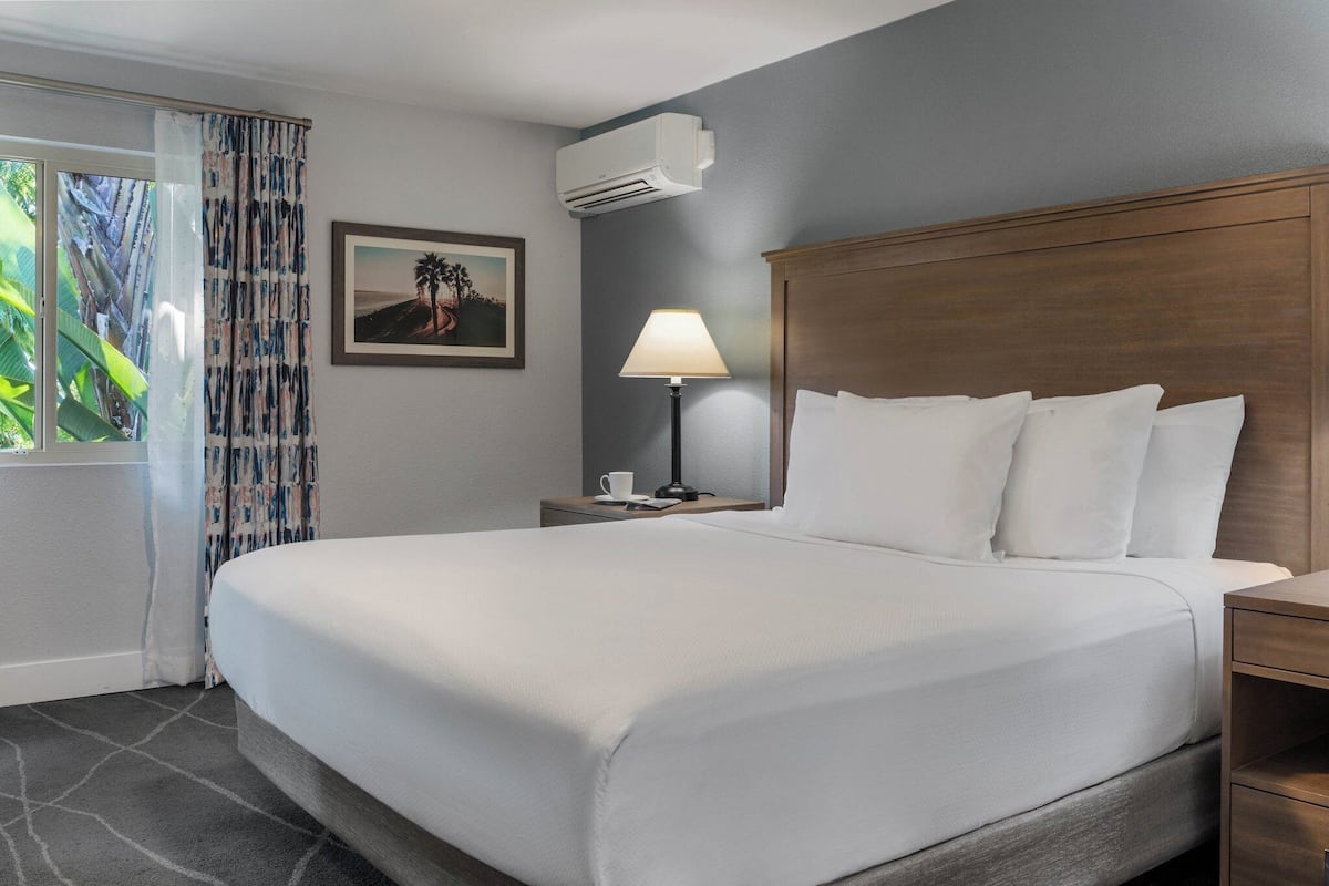 A hotel room with a large bed, a nightstand with a lamp, a framed picture on the wall, and an air conditioning unit.