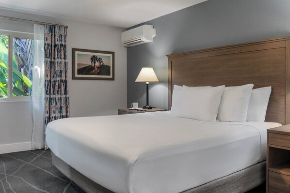 Quarto de hotel com uma cama grande, uma mesa de cabeceira com abajur, um quadro na parede e um aparelho de ar condicionado.
