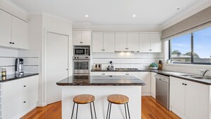 Una cocina moderna con armarios blancos, una isla central con taburetes, electrodomésticos de acero inoxidable y vistas a través del ventanal.