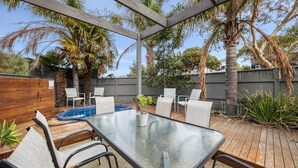 Un patio al aire libre con una mesa de vidrio, sillas blancas, una piscina pequeña y diversas plantas.