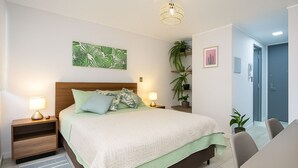 A neatly arranged bedroom with a bed, bedside tables, a lamp, a hanging plant, and a door. - Apartment w/private parking, steps from Teatro Caupolicán and minutes from Movistar. (Santiago)