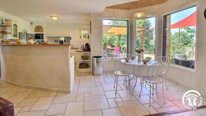 A modern kitchen with a central island, a dining area with white chairs, and large windows offering a view of the outdoors. - Saint Co - SARZEAU - Gîte (SARZEAU)