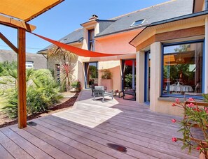 Een modern huis met een houten terras, zitgedeelte buiten en een groot raam met uitzicht op de tuin.