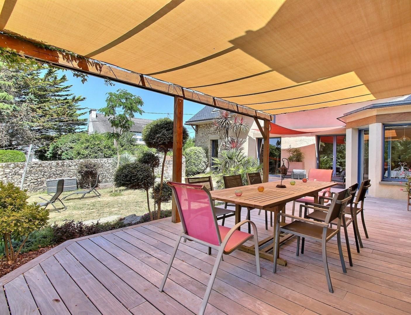 An outdoor patio with a wooden table and chairs, a pergola providing shade, and a stone wall in the background.